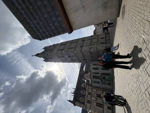       Two travellers posing in front of the tall medieval Belfry tower on a cobbled square under dramatic clouds
  