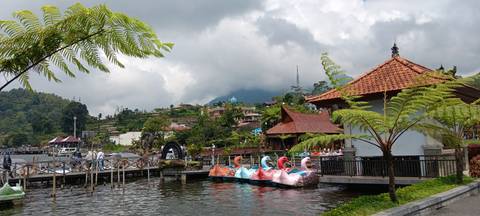       Lakeside scene with swan paddle boats, Balinese pavilion, and misty mountain backdrop.
  