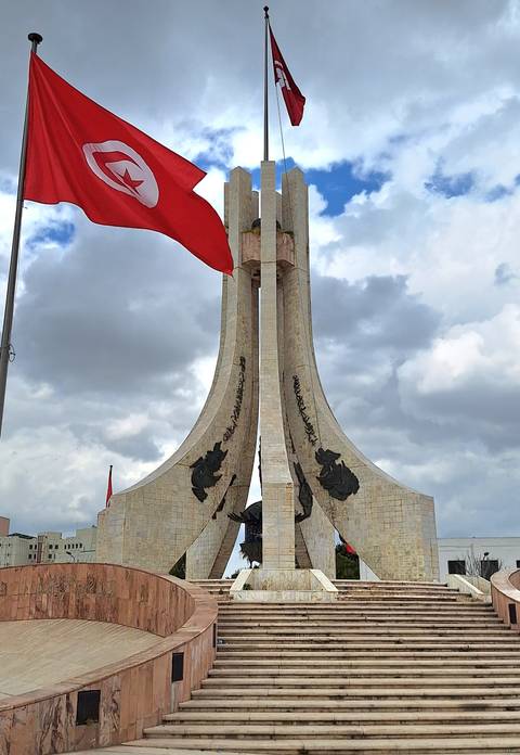       Tall modernist monument with Tunisian flag waving against a cloudy sky.
  