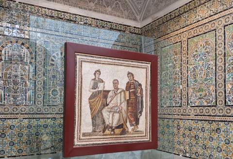       Ancient Roman mosaic displayed on a stand within a richly tiled gallery.
  