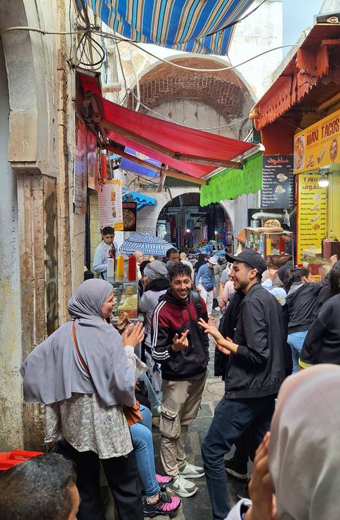       Crowded medina alley with vibrant signs, food stalls and animated locals chatting.
  