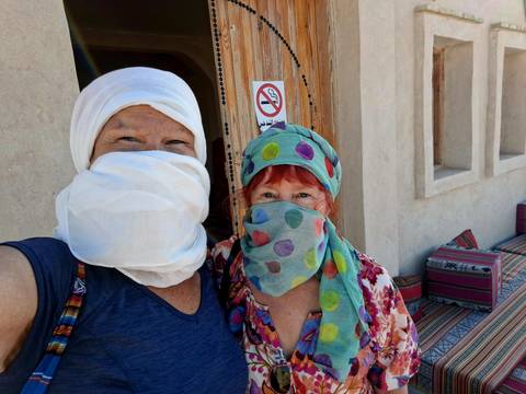       Two women wearing colorful desert scarves pose for a cheerful selfie outside a mud-brick building.
  