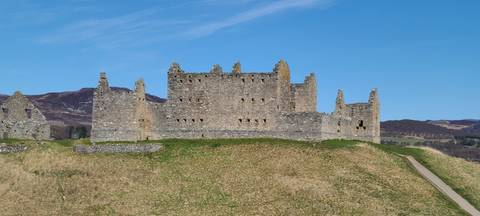       Ruthven Barracks stone ruins stand isolated on a grassy mound with mountains behind.
  