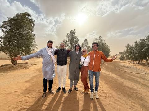       Five travellers pose cheerfully on a windy desert road lined with trees and blowing sand
  