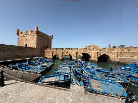       Blue fishing boats cluster in Essaouira’s historic stone harbor beside a fortified tower
  