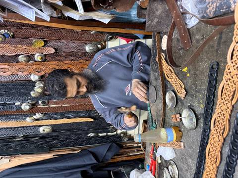       A craftsman hammers leather pieces at a cluttered workshop stall filled with belts and tools
  