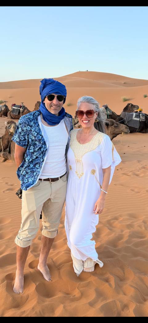       A smiling couple in traditional attire stand before resting camels on desert sand
  