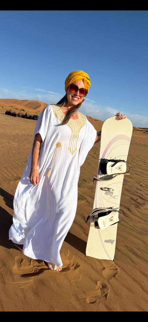       A woman in white dress holds a sandboard on golden dunes under blue sky
  