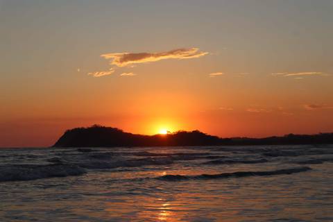       Fiery orange sun setting behind a headland, lighting up gentle waves on a tranquil beach.
  