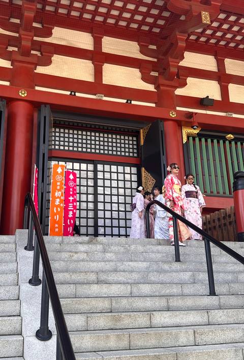       Women wearing colorful kimonos descend temple steps with vermillion columns and decorative lattice doors.
  