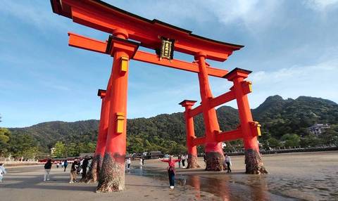       Iconic vermillion Great Torii of Miyajima rising from sandy tidal flats with forested hills behind.
  
