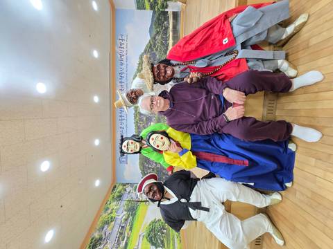       Tourist seated with masked performers wearing colorful traditional Korean costumes inside a cultural center.
  