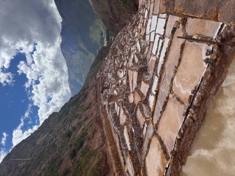       Terraced salt evaporation ponds of Maras gleaming under patchy clouds with Andean peaks behind.
  