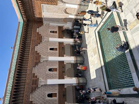       Intricately tiled courtyard and pool of a historic medersa with visitors admiring the architecture.
  