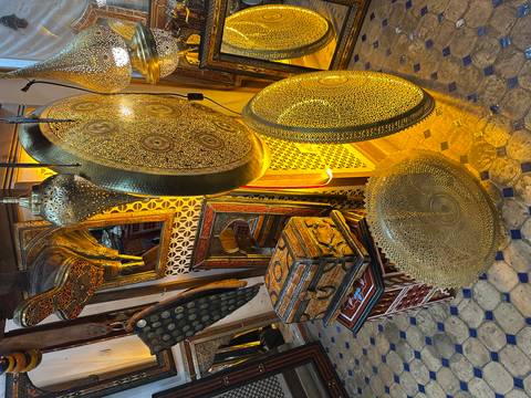       An ornate Moroccan shop interior displaying large hammered-brass trays, lanterns and painted wooden furniture.
  