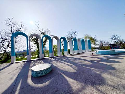       Open plaza featuring a row of turquoise-and-white arches casting long shadows under a clear blue sky.
  
