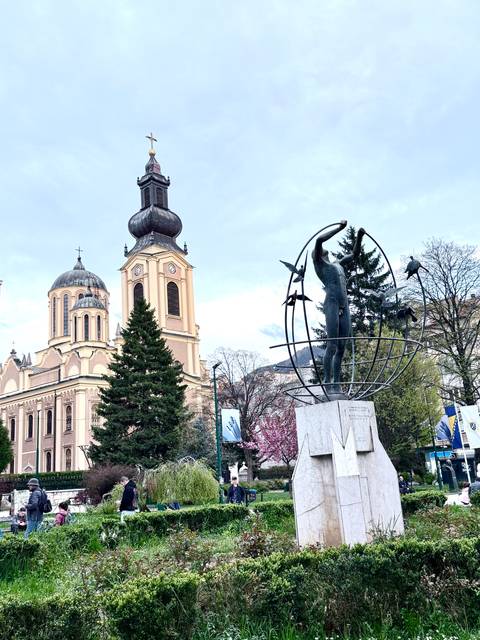       Pink-toned church tower beside a modern globe statue with birds in an urban square framed by trees.
  