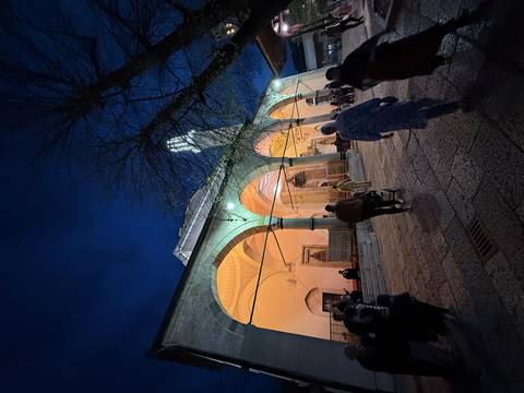       Evening scene of a mosque courtyard lit with warm lights as visitors walk beneath stone arches.
  