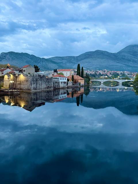       Calm river reflecting stone walls, red-roofed houses and an arched bridge with mountains in the background.
  