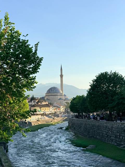       Panoramic view of a domed mosque and tall minaret rising above a historic town with snowy peaks behind.
  