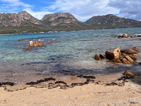       Clear turquoise water lapping sandy shore with forested granite peaks rising behind under patchy clouds.
  