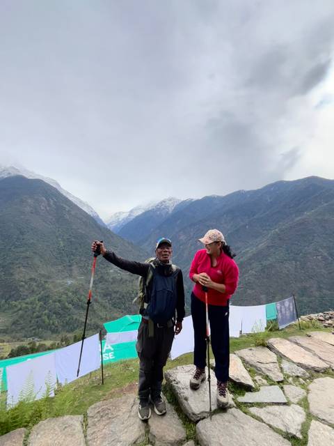       Two trekkers with poles smile on a green valley ridge backed by snow-capped Himalayan mountains.
  