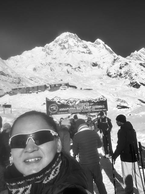       Black-and-white snapshot of trekkers gathered at Annapurna Base Camp sign with snowy slopes behind.
  