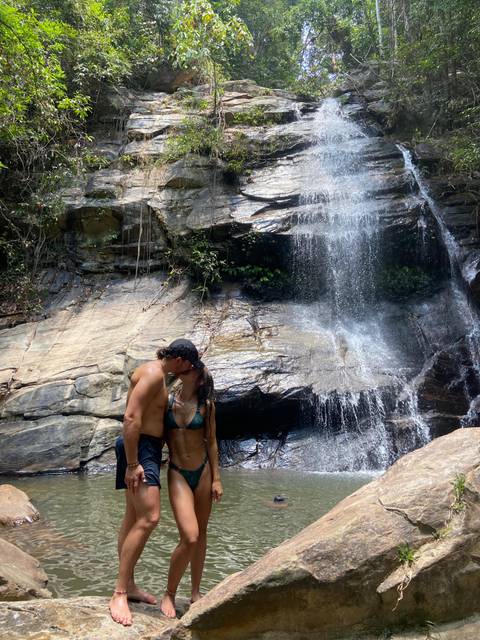       Couple in swimwear shares a kiss beneath a tropical waterfall cascading over smooth rock.
  