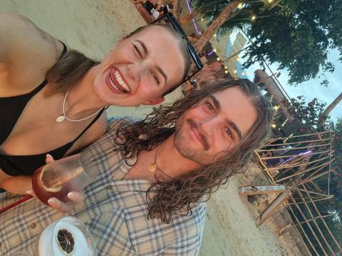       Smiling young friends take a selfie with drinks at a beach bar decorated with lights and bamboo.
  