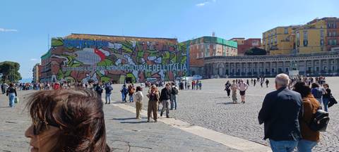       Large open square in Naples with colourful mural-clad building, crowds of tourists and locals under bright skies.
  