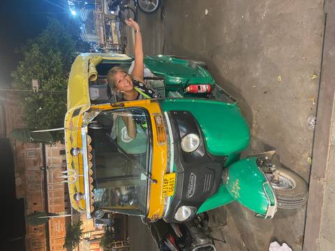       Smiling traveler leans out of a green and yellow auto-rickshaw at night on a busy Indian street.
  