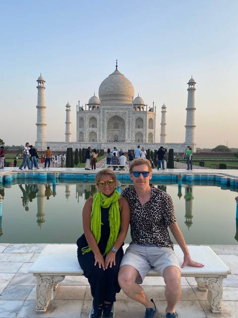       Couple seated beside a reflecting pool with the Taj Mahal rising majestically behind them at dusk.
  