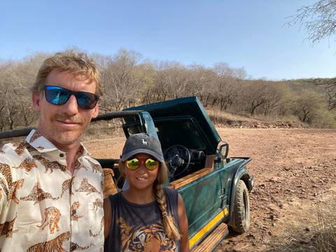       Selfie of two travelers beside an open-top jeep in a dry forest landscape during a safari.
  
