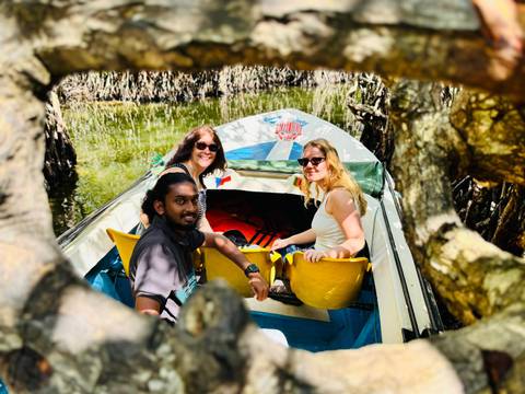       Three travelers smile from a small boat paddling through lush mangrove tunnels framed by twisted branches.
  