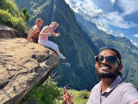       Trio of hikers sit and give a thumbs-up on a dramatic cliff ledge overlooking green valleys.
  