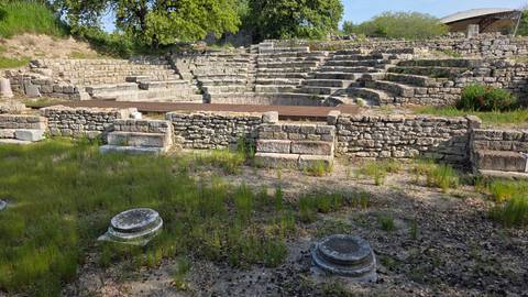       Stone-built semicircular ancient theatre with tiered seating surrounded by low grass and wildflowers.
  