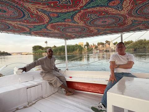       Two men relax on a traditional felucca boat gliding along the Nile at dusk with riverbanks behind.
  