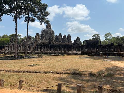       Wide view of the stone towers and galleries of Bayon Temple surrounded by jungle under blue skies.
  