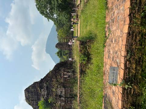       Tourists walk among grassy ruins with a distant mountain rising beyond My Son Sanctuary.
  