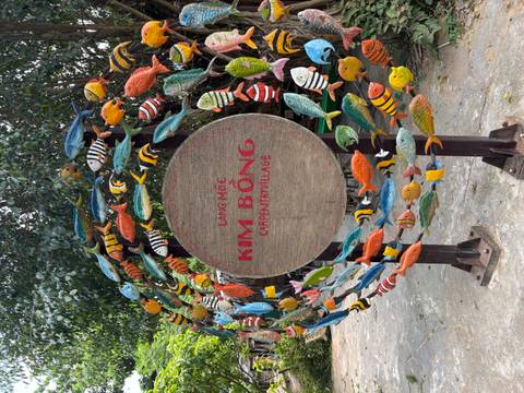       Colorful fish carvings form a round sign reading 'Kim Bồng Carpentry Village' in a leafy courtyard.
  