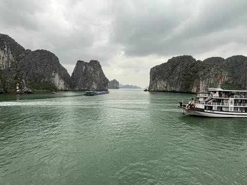       Boats cruise between dramatic limestone towers in green waters of Halong Bay under cloudy skies.
  
