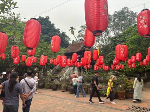       Dozens of red lanterns hang above visitors exploring an outdoor temple courtyard surrounded by greenery.
  