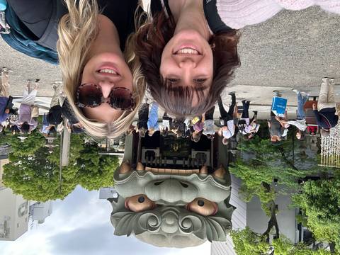       Two travelers smile before giant open-mouthed lion head shrine as crowds wander nearby.
  