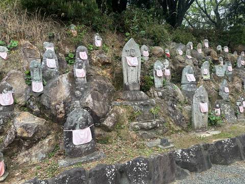       Row of stone Jizo statues on rocky hillside each wearing pink cloth bibs.
  