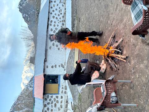       Two men warming by a tall orange campfire flame outside a rustic lodge.
  