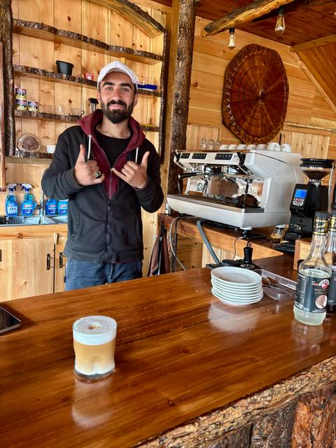       Barista standing behind a wooden coffee counter with a professional espresso machine.
  