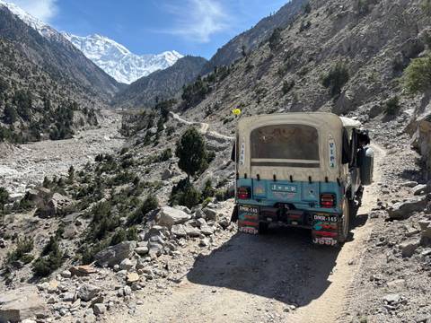       Open-back jeep driving along a rugged mountain track toward snowy peaks.
  