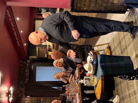       Man presenting traditional food to a seated group in a cozy whisky bar with maroon walls.
  