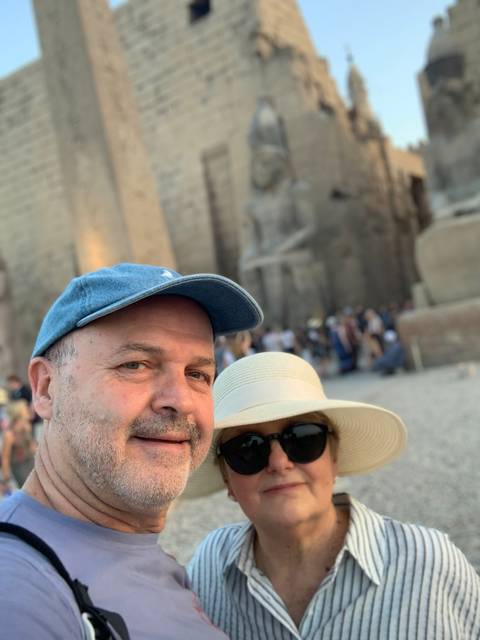       Close-up selfie of a couple wearing hat and cap with ancient statues blurred behind.
  