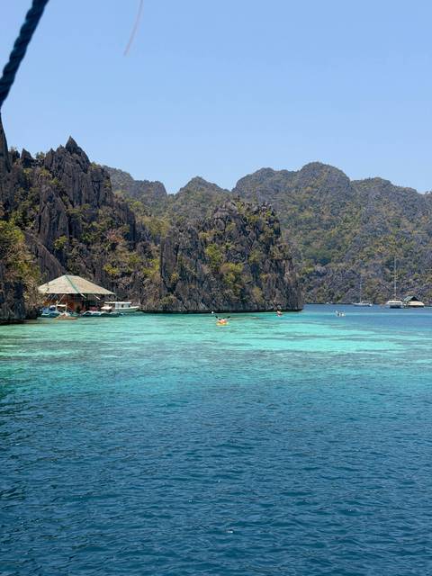       Turquoise lagoon surrounded by dramatic limestone cliffs with kayakers and a floating hut in Palawan.
  